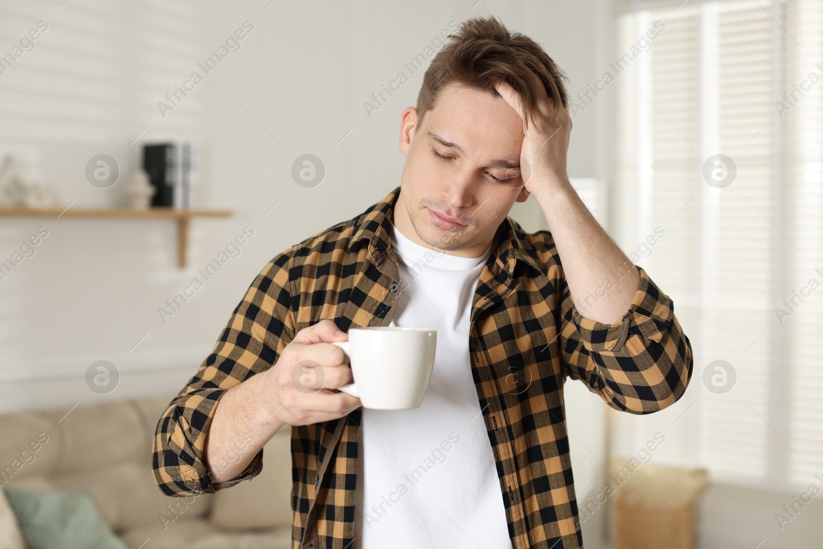 Photo of Unhappy young man with cup of coffee suffering from hangover at home