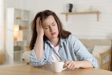 Photo of Suffering from hangover. Unhappy woman with cup of coffee sitting at wooden table indoors