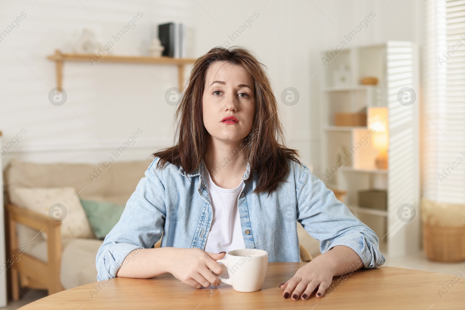 Photo of Suffering from hangover. Unhappy woman with cup of coffee sitting at wooden table indoors