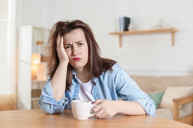 Suffering from hangover. Unhappy woman with cup of coffee sitting at wooden table indoors