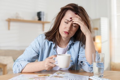 Suffering from hangover. Unhappy woman with cup of coffee sitting at wooden table indoors