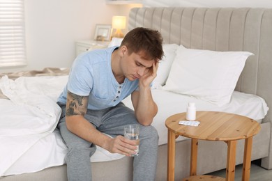 Photo of Unhappy young man with glass of water suffering from hangover on bed at home