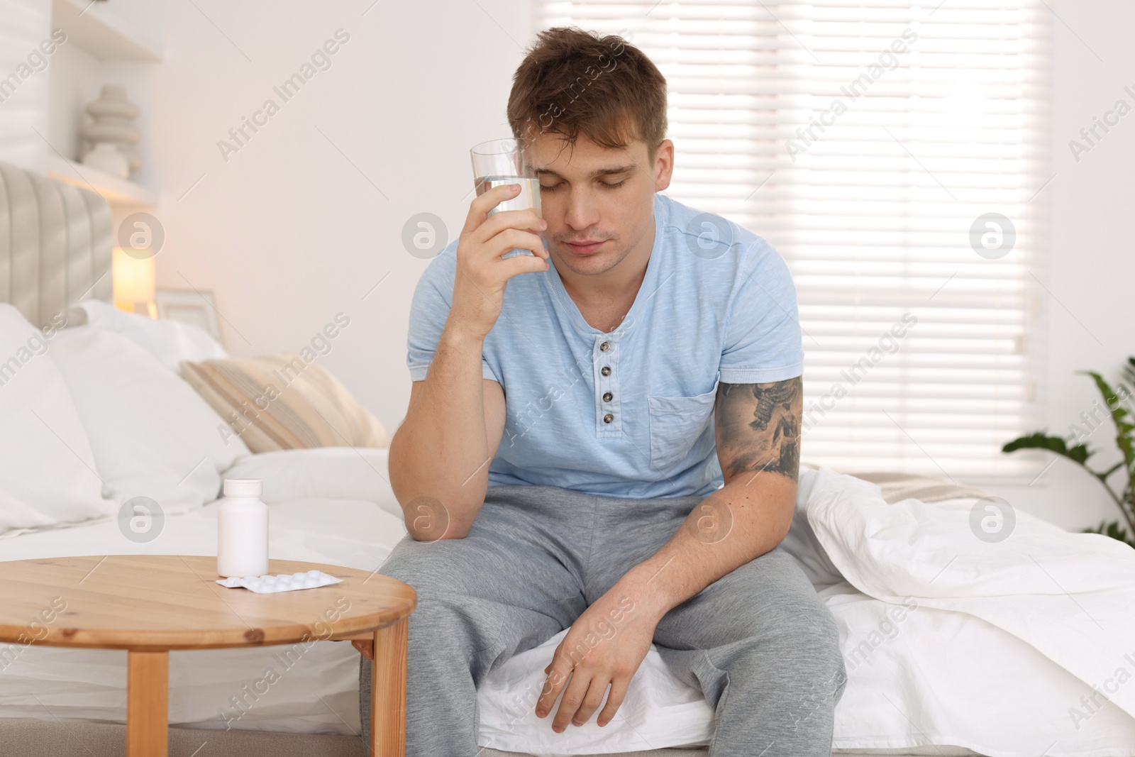 Photo of Unhappy young man with glass of water suffering from hangover on bed at home