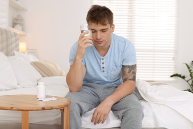 Unhappy young man with glass of water suffering from hangover on bed at home