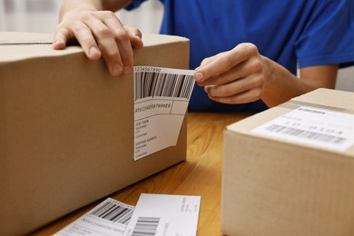 Photo of Man sticking shipping label with barcode on parcel at wooden table indoors, closeup