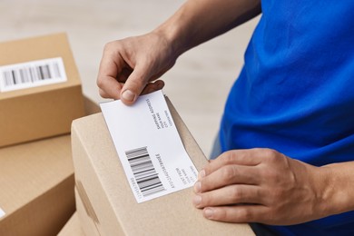 Man sticking shipping label with barcode on parcel indoors, closeup