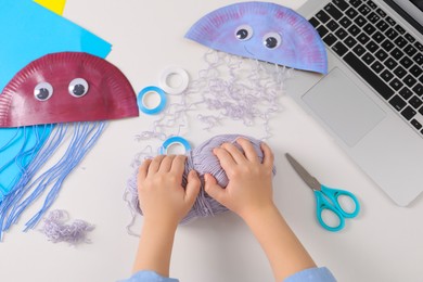 Photo of Little girl making handmade jellyfish by craft tutorial video at white table, top view. Child creativity