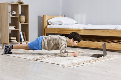 Photo of Little boy doing plank exercise near laptop at home. Morning routine