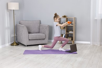 Photo of Little girl exercising with dumbbells near laptop at home. Morning routine