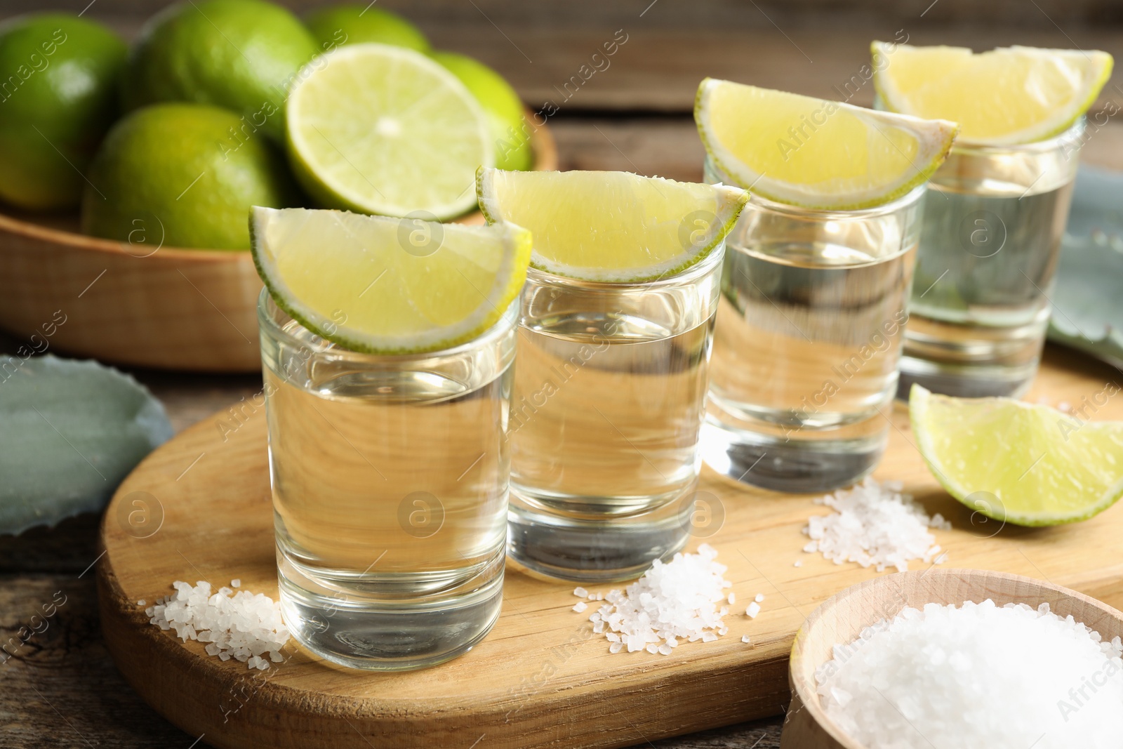 Photo of Tequila shots with lime slices, salt and agave leaves on wooden table, closeup