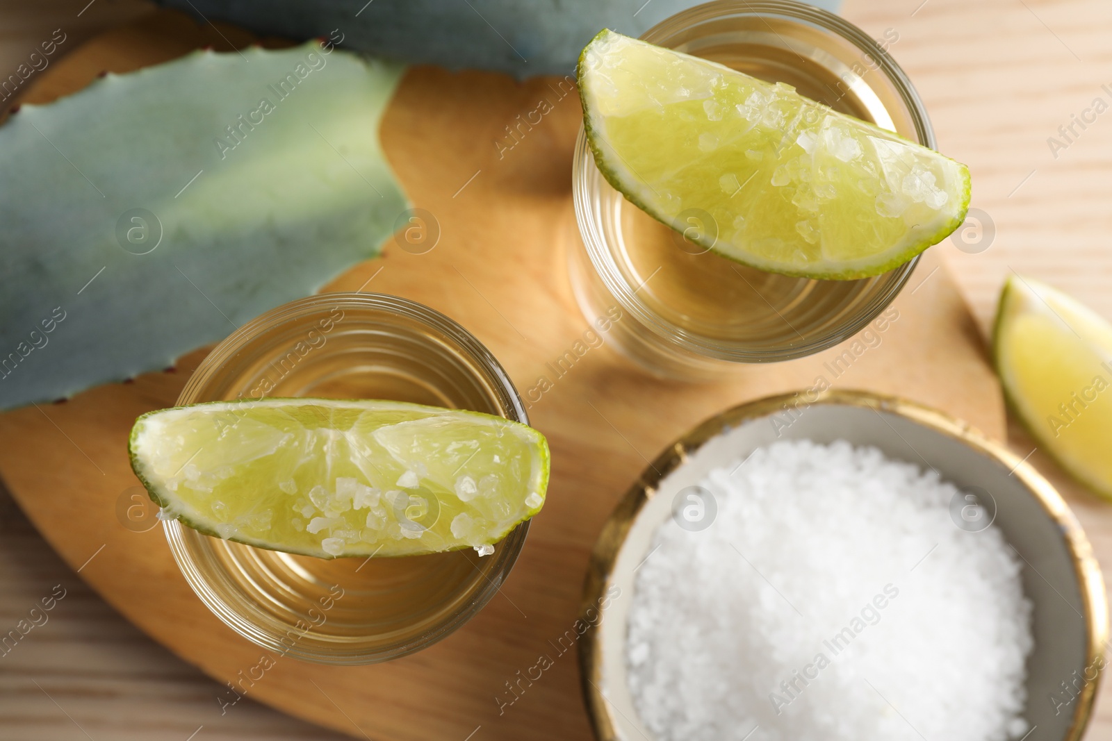 Photo of Tequila shots with lime slices, salt and agave leaves on wooden table, flat lay