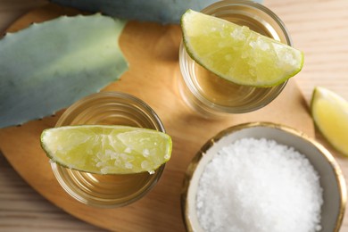 Tequila shots with lime slices, salt and agave leaves on wooden table, flat lay