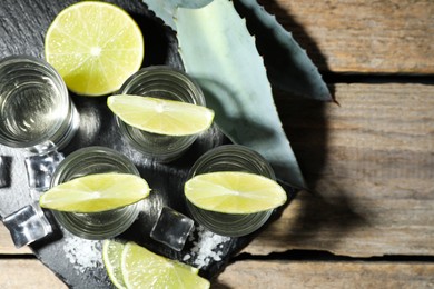 Tequila shots with lime slices, salt, ice cubes and agave leaves on wooden table, flat lay