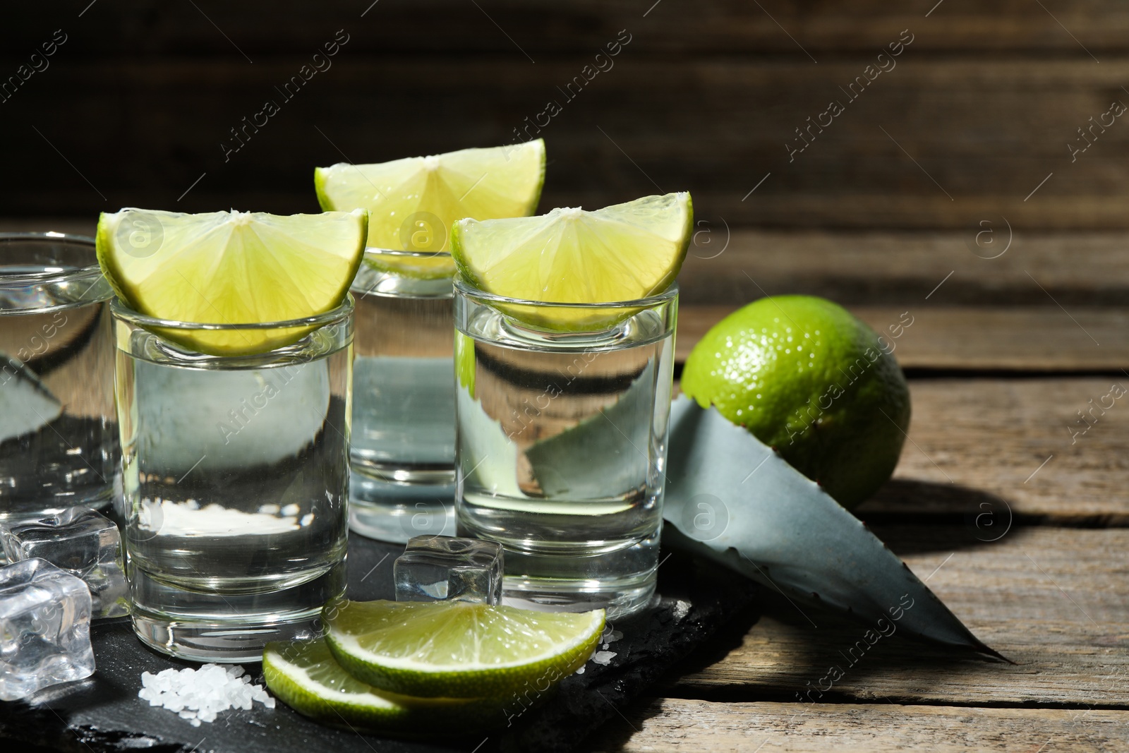 Photo of Tequila shots with lime slices, salt, ice cubes and agave leaf on wooden table, closeup