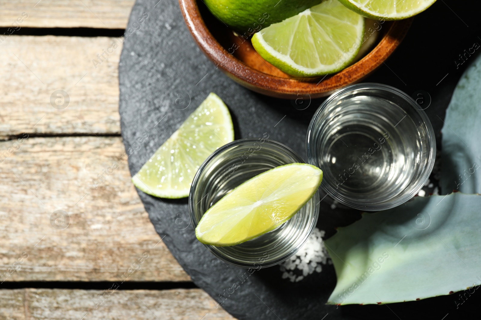 Photo of Tequila shots with lime slices, salt and agave leaves on wooden table, flat lay