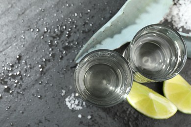 Photo of Tequila shots, slices of lime, salt and agave leaves on wet grey table, flat lay. Space for text