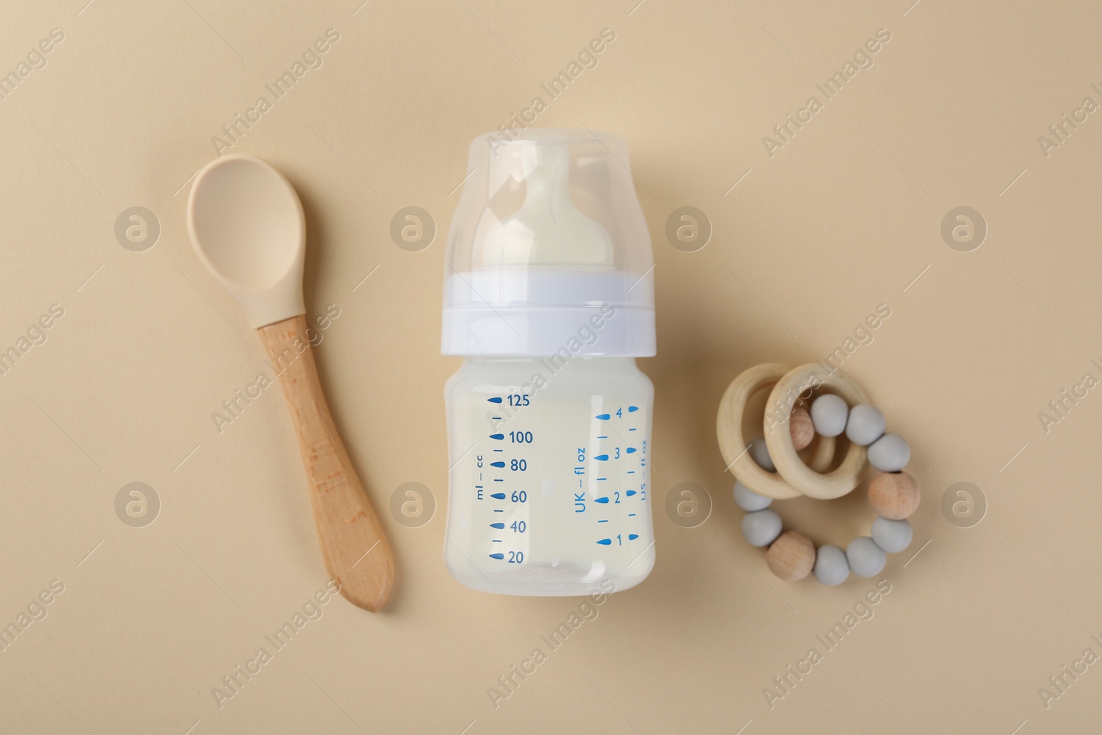 Photo of Feeding bottle with milk, spoon and teether on beige background, flat lay