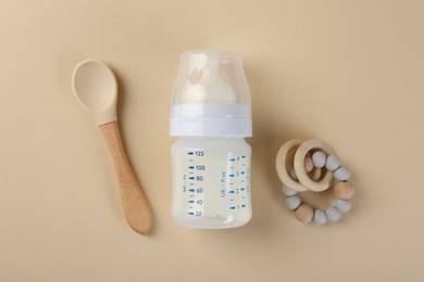 Feeding bottle with milk, spoon and teether on beige background, flat lay