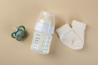 Feeding bottle with milk, baby socks and pacifier on beige background, flat lay