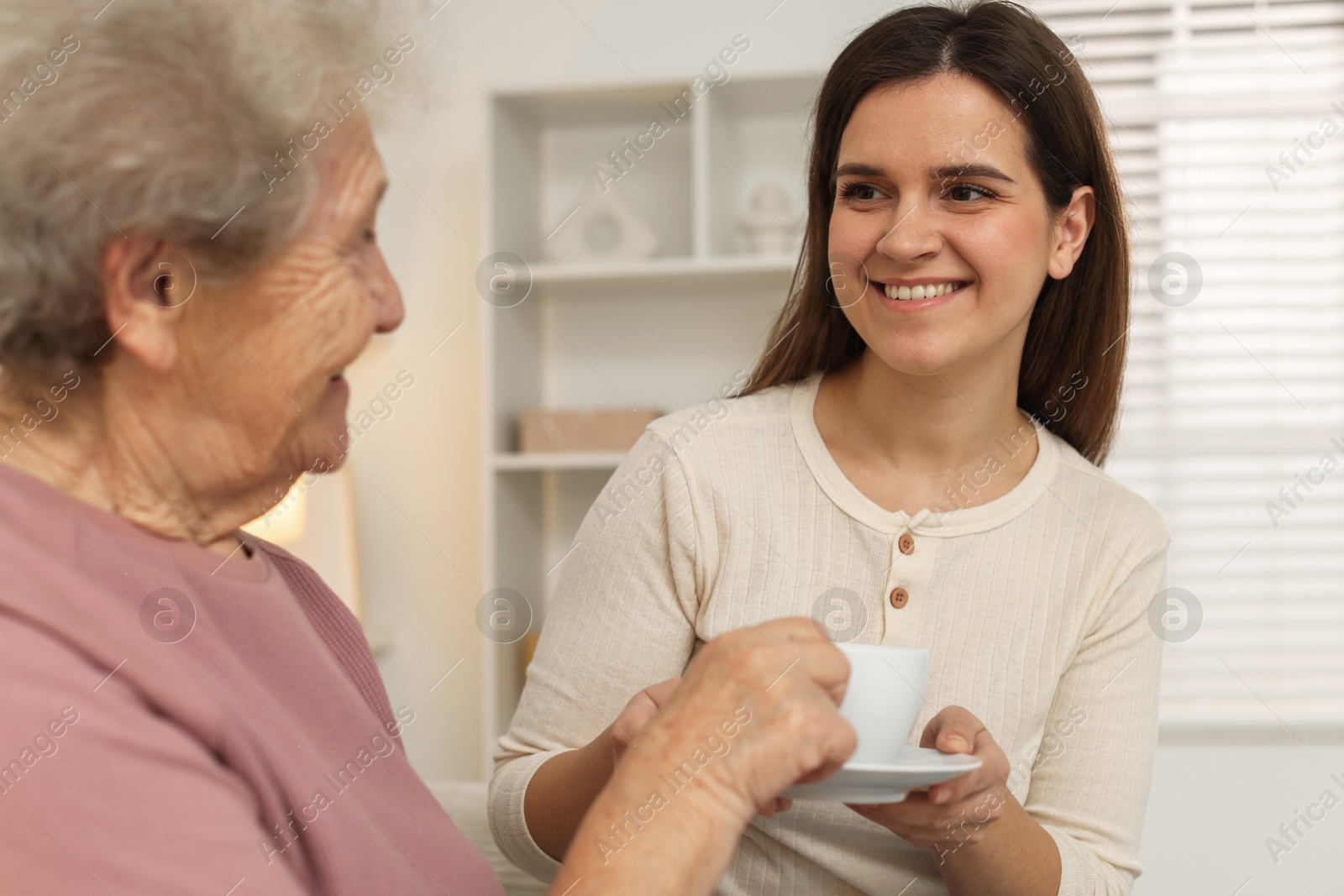 Photo of Granddaughter giving hot drink to her grandmother at home. Elderly care