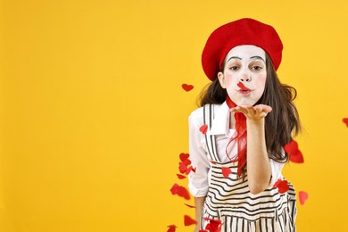 Photo of Cute girl dressed like mime blowing out confetti in shape of hearts on yellow background, space for text. Surprise party