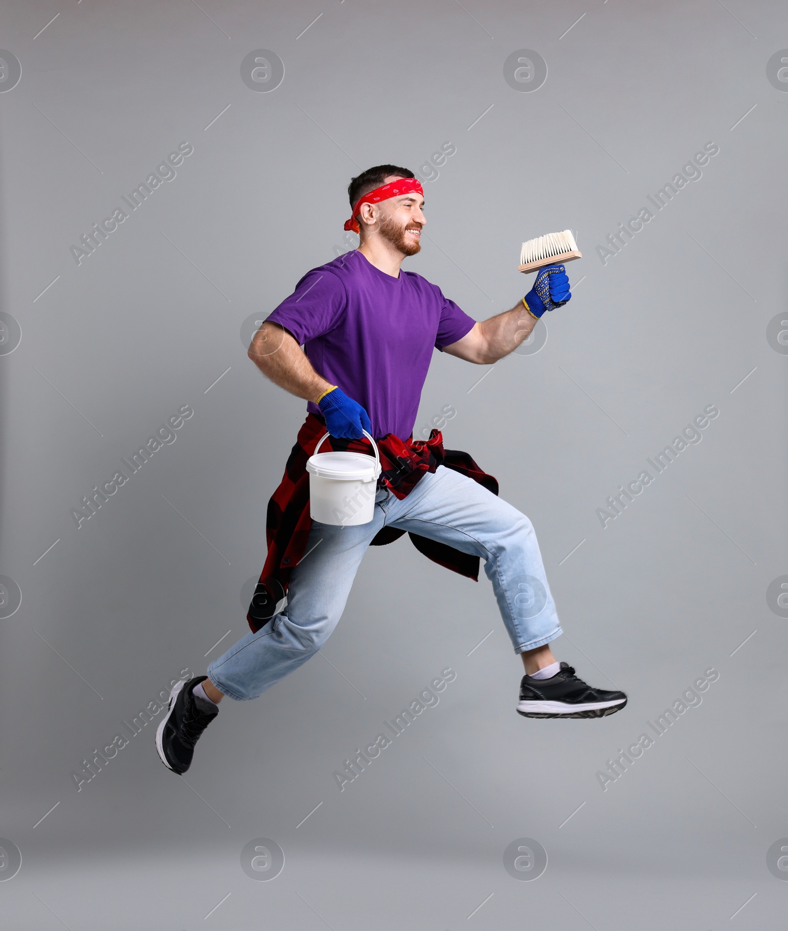 Photo of Man with brush and bucket of paint jumping on light grey background