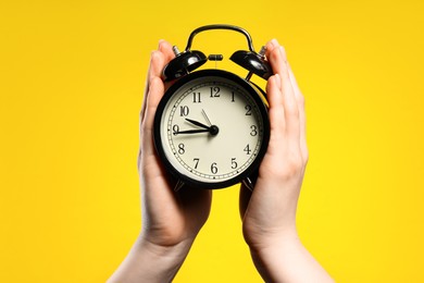 Photo of Woman with black alarm clock on yellow background, closeup