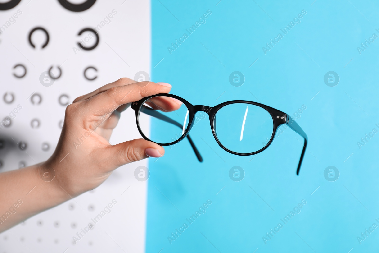 Photo of Woman holding glasses against vision test chart on light blue background, closeup