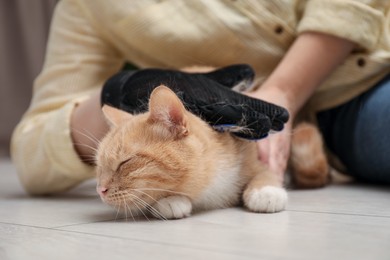 Photo of Woman brushing cat's hair with glove on floor at home, closeup. Pet grooming