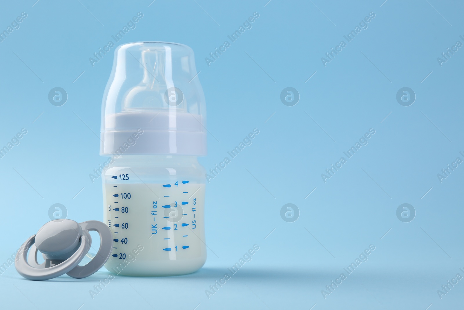 Photo of Feeding bottle with milk and pacifier on light blue background, space for text