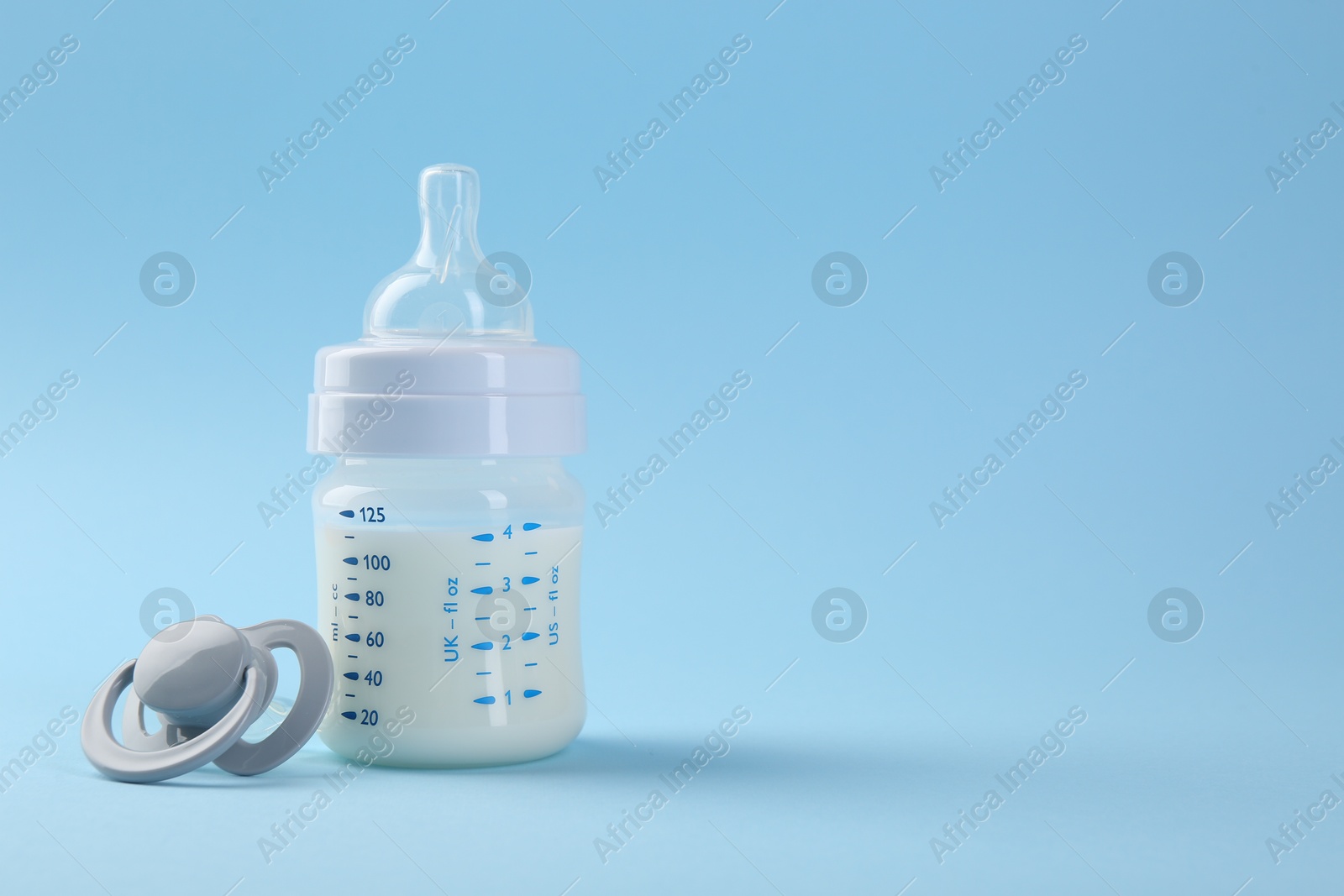 Photo of Feeding bottle with milk and pacifier on light blue background, space for text