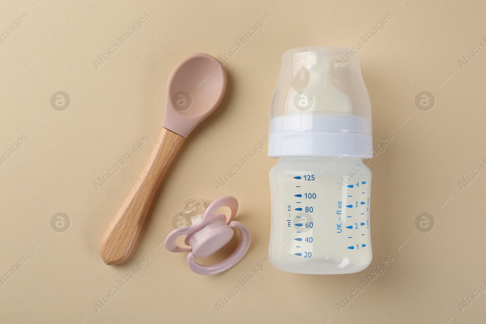 Photo of Feeding bottle with milk, spoon and pacifier on beige background, flat lay