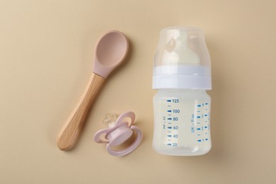 Feeding bottle with milk, spoon and pacifier on beige background, flat lay