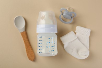 Feeding bottle with milk, spoon, baby socks and pacifier on beige background, flat lay