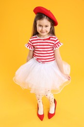 Photo of Stylish little girl wearing oversized high heeled shoes on orange background