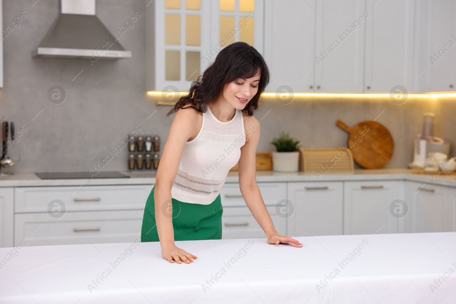 Photo of Young woman putting white tablecloth on table in kitchen