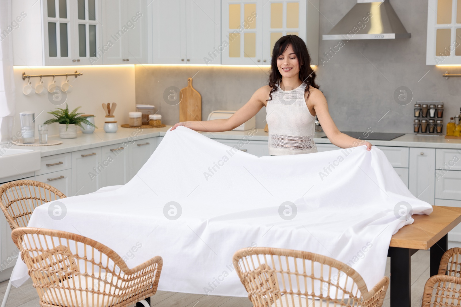 Photo of Young woman putting white tablecloth on table in kitchen