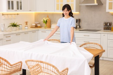 Photo of Young woman putting white tablecloth on table in kitchen