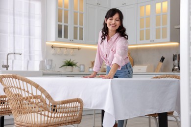 Photo of Young woman putting white tablecloth on table in kitchen