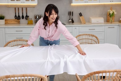 Photo of Young woman putting white tablecloth on table in kitchen