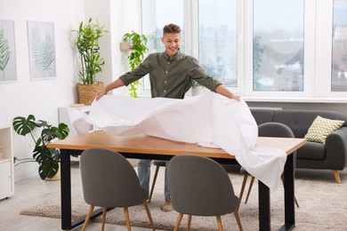 Photo of Young man putting white tablecloth on table at home