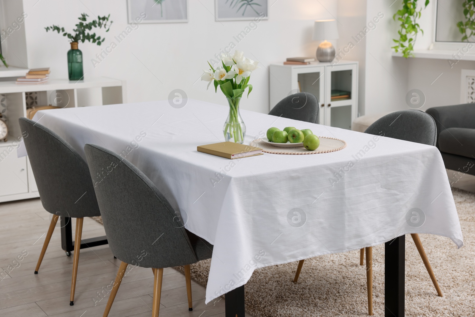 Photo of Table with white tablecloth, apples, flowers in vase and chairs in stylish room. Interior design