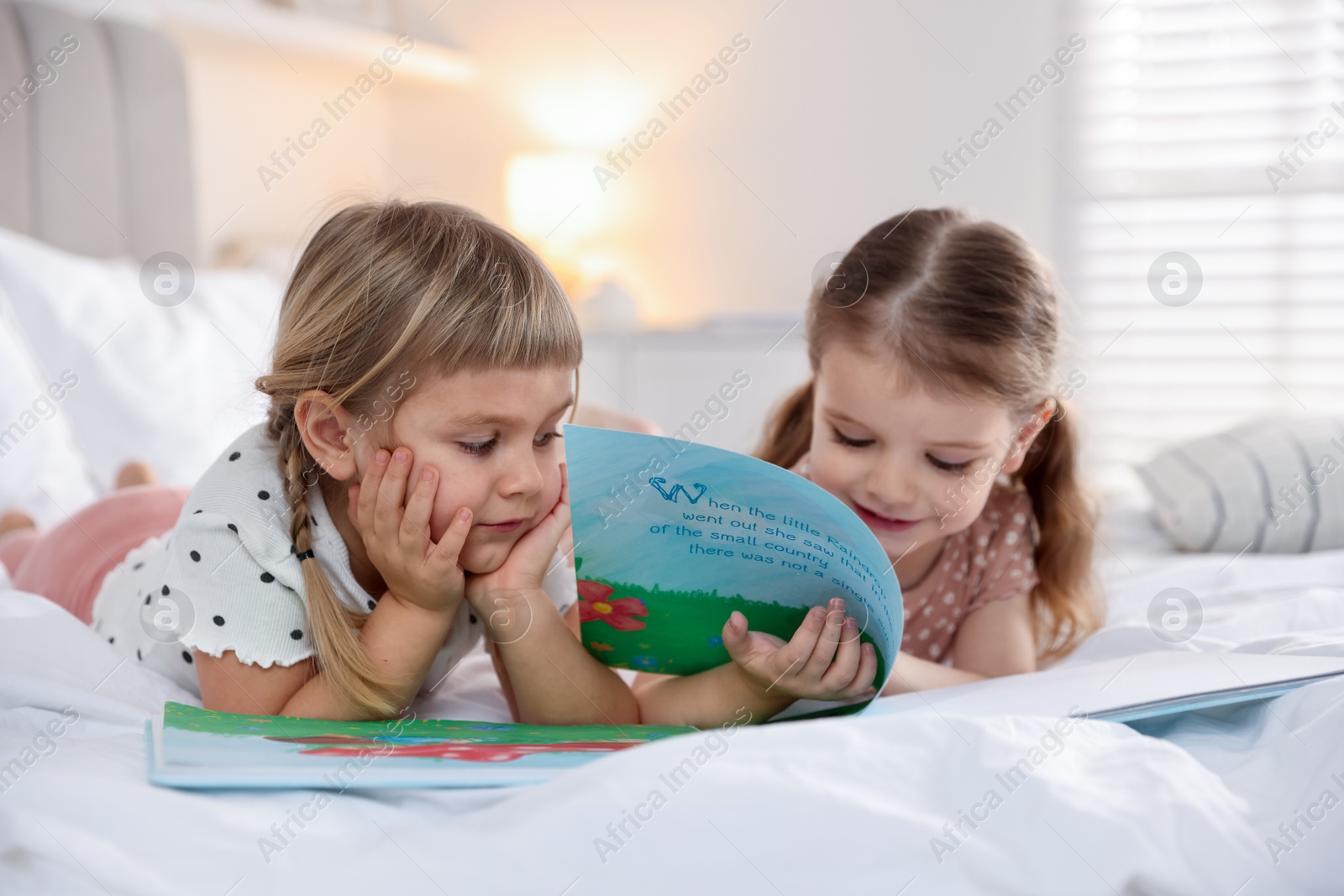 Photo of Cute little sisters reading book together on bed at home