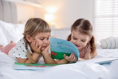 Cute little sisters reading book together on bed at home