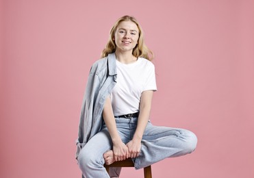 Smiling woman in stylish jeans sitting on stool against pink background