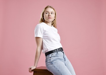 Photo of Woman in stylish jeans sitting on stool against pink background, low angle view