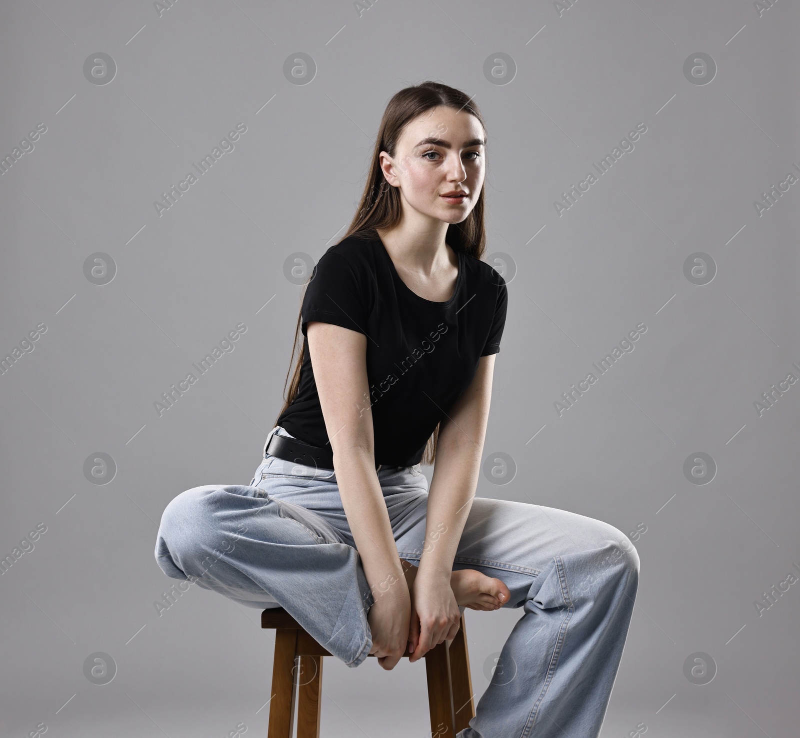 Photo of Beautiful young woman in stylish jeans sitting on stool against grey background