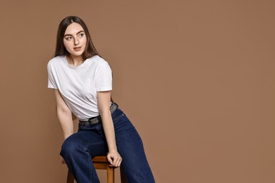 Photo of Woman in stylish jeans sitting on stool against brown background. Space for text