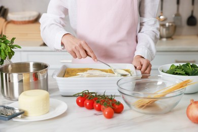 Woman spreading bechamel sauce onto spinach lasagna at marble table indoors, closeup