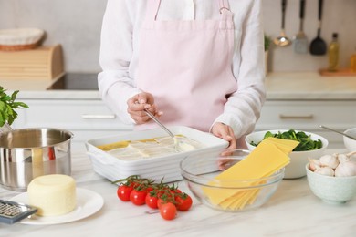 Photo of Woman spreading bechamel sauce onto spinach lasagna at marble table indoors, closeup